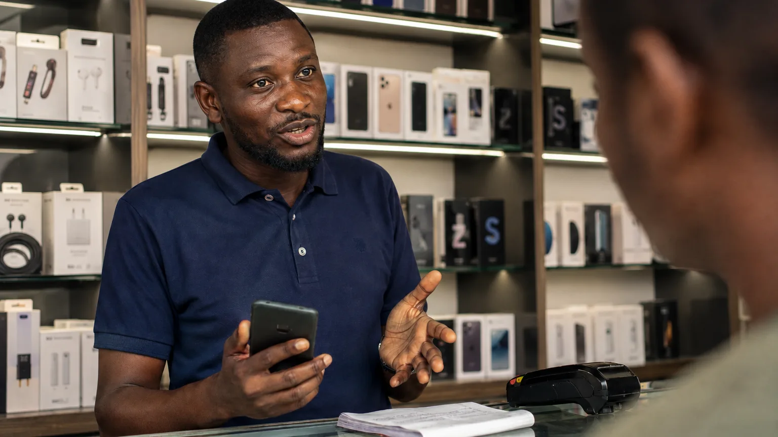 A phone seller behind the glass counter of his shop, phone in hand, explaining a sale to a customer, with lit shelves of boxed phones and accessories behind him.